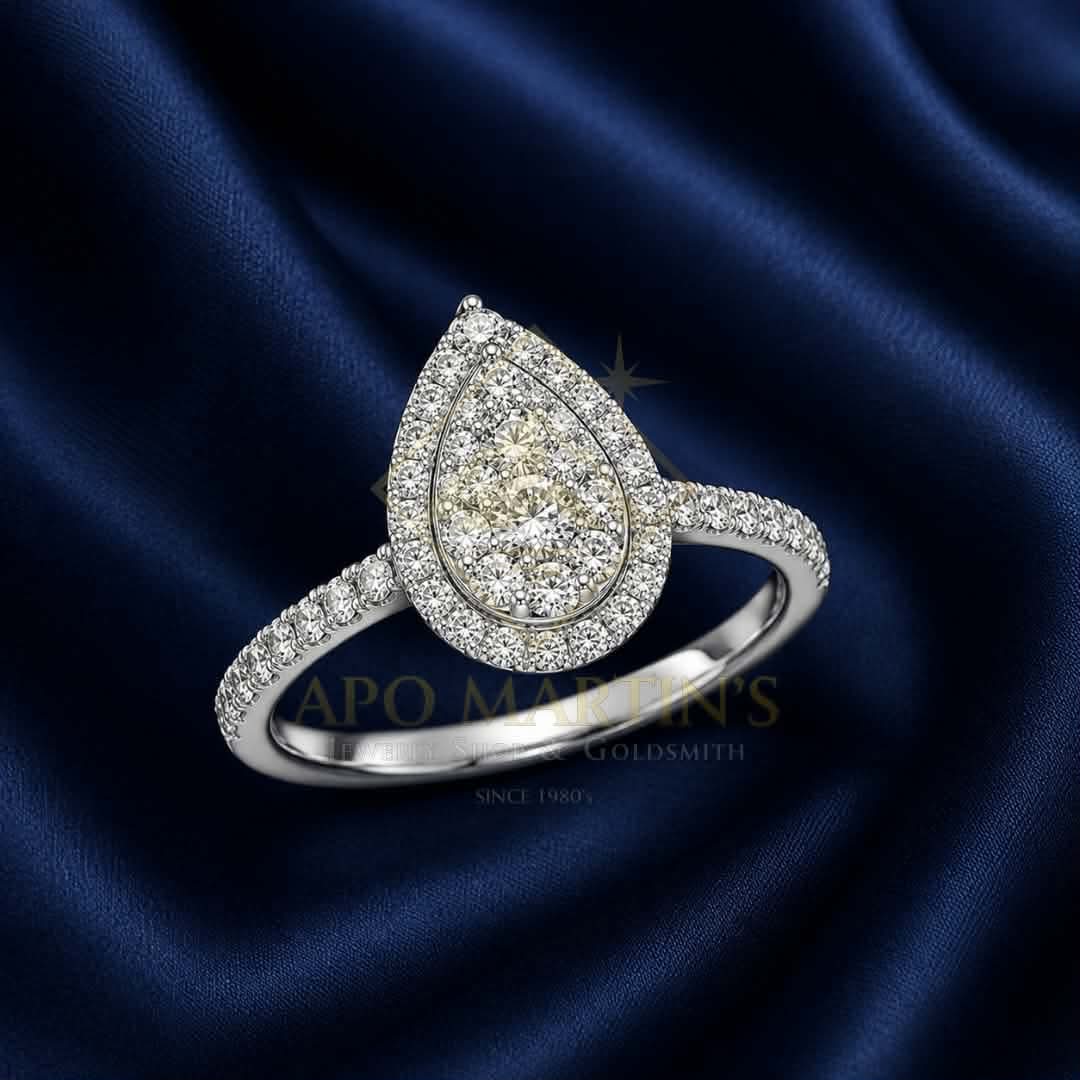 A teardrop-shaped diamond ring with a halo setting and pavé band, displayed on a dark blue satin fabric background. A watermark for 'Apo Martin's Goldsmith' is visible across the center.