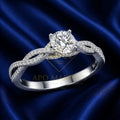 A silver-toned twist band engagement ring with a round-cut center stone and pavé-set accent stones, displayed on a draped dark blue satin fabric. A watermark for 'APO MARTIN'S' is visible across the center.