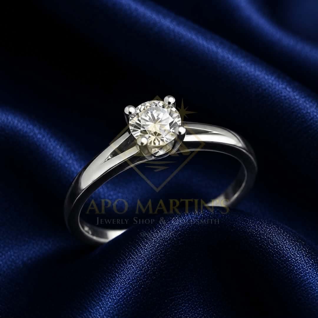 A silver solitaire engagement ring with a round-cut diamond set in a four-prong setting, displayed on a draped dark blue satin fabric background. A watermark for 'APO MARTINS Jewelry Shop & Gemsmith' is visible in the center.