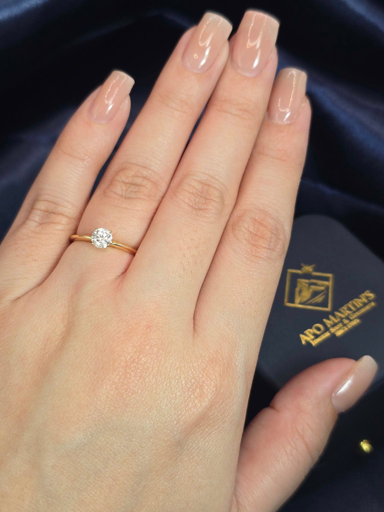A close-up of a hand wearing a yellow gold engagement ring with a round-cut diamond. The hand has a glossy nude manicure and is resting on a dark navy blue fabric surface next to a dark blue jewelry box with the brand name 'Ard Martin's'.