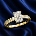A yellow gold solitaire engagement ring with a cushion-cut clear center stone and pavé-set accent stones on the band, displayed on a dark blue satin fabric background.