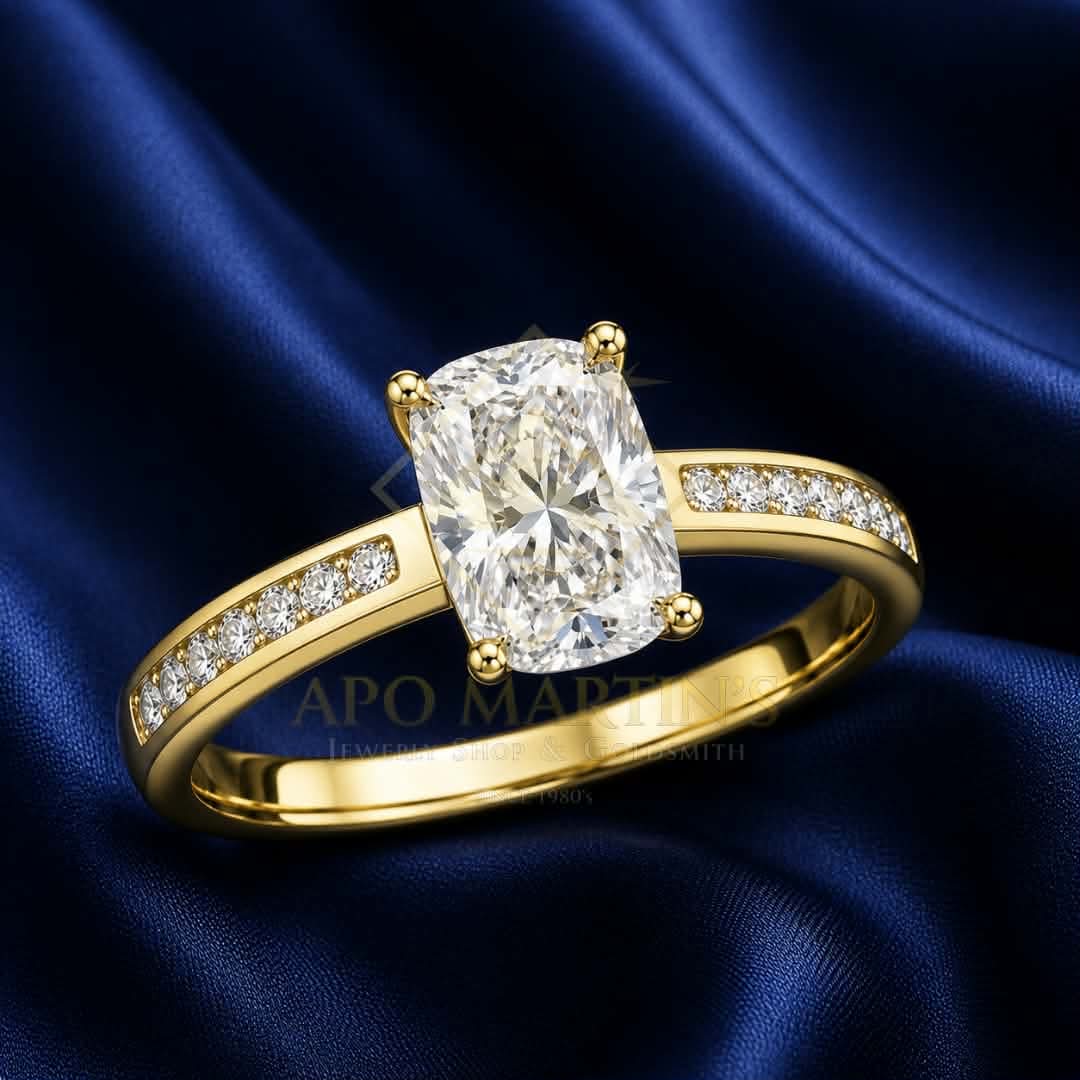 A yellow gold solitaire engagement ring with a cushion-cut clear center stone and pavé-set accent stones on the band, displayed on a dark blue satin fabric background.