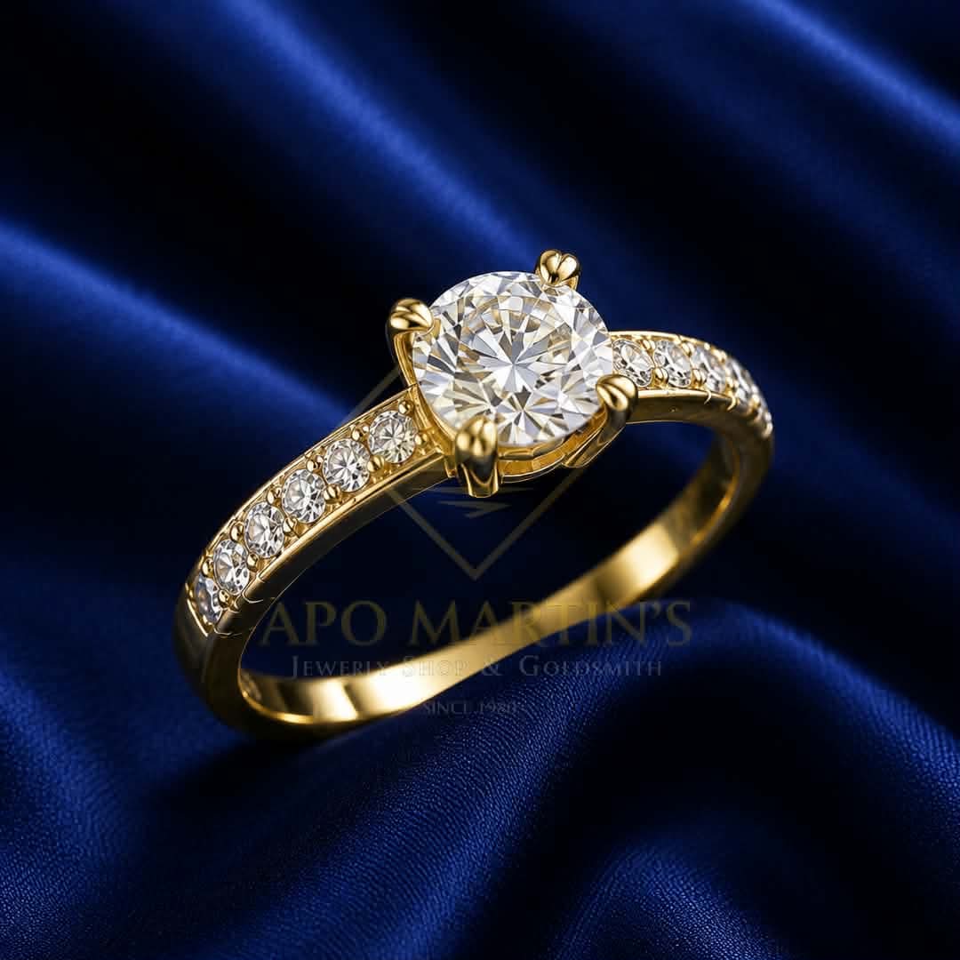 A yellow gold solitaire engagement ring with a round-cut diamond center stone and pavé-set diamonds along the band, displayed on a dark blue satin fabric background.