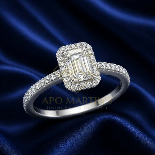 An emerald-cut halo ring with a silver-toned band paved with small diamonds, displayed on a dark blue satin fabric background. A watermark for 'APO MARTINS Jewelry Smith' is visible across the center.