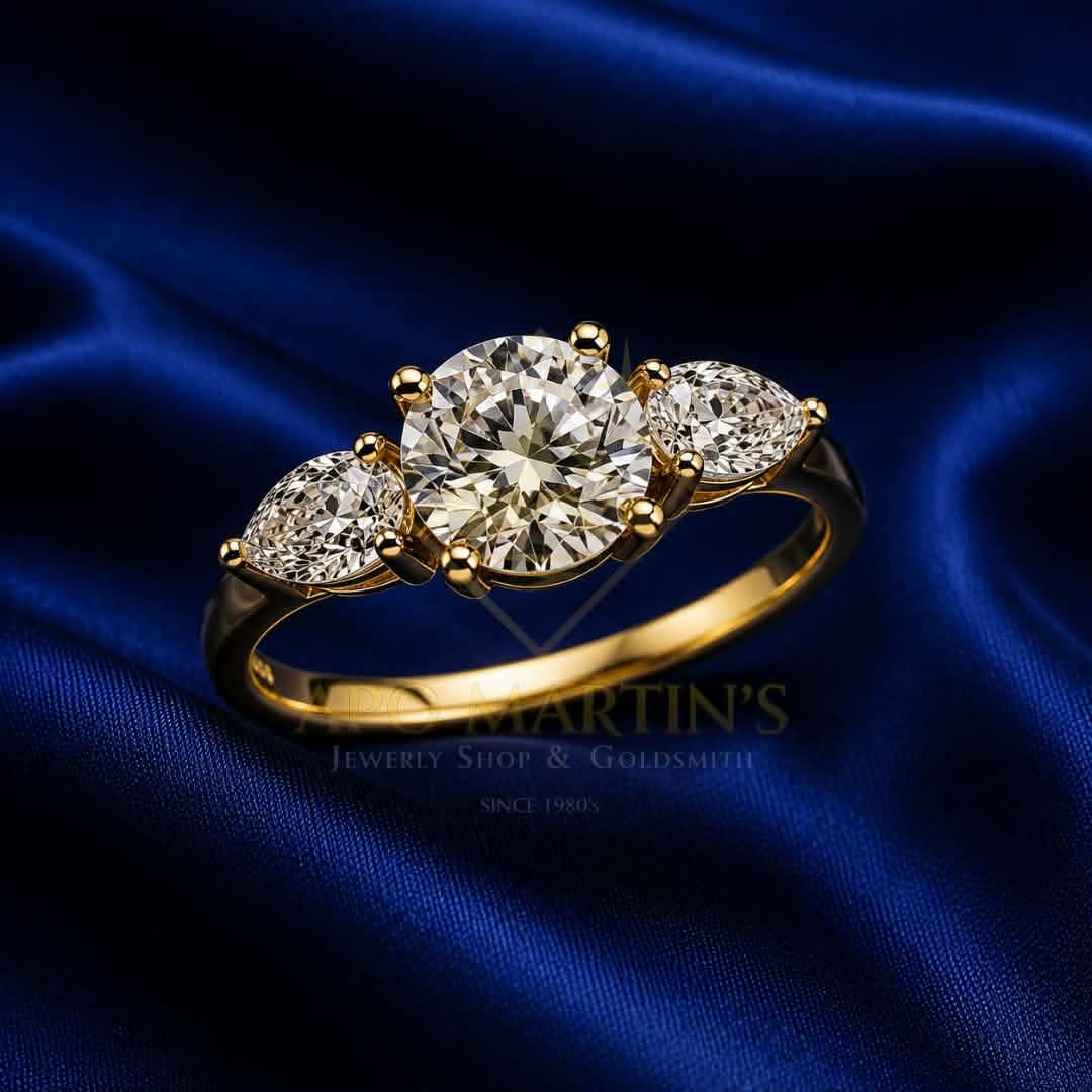 A yellow gold three-stone engagement ring with a round center diamond and marquise-cut side diamonds, displayed on a dark blue satin fabric background. A faint watermark for 'Martin's Jewelry Shop & Goldsmith' is visible on the image.