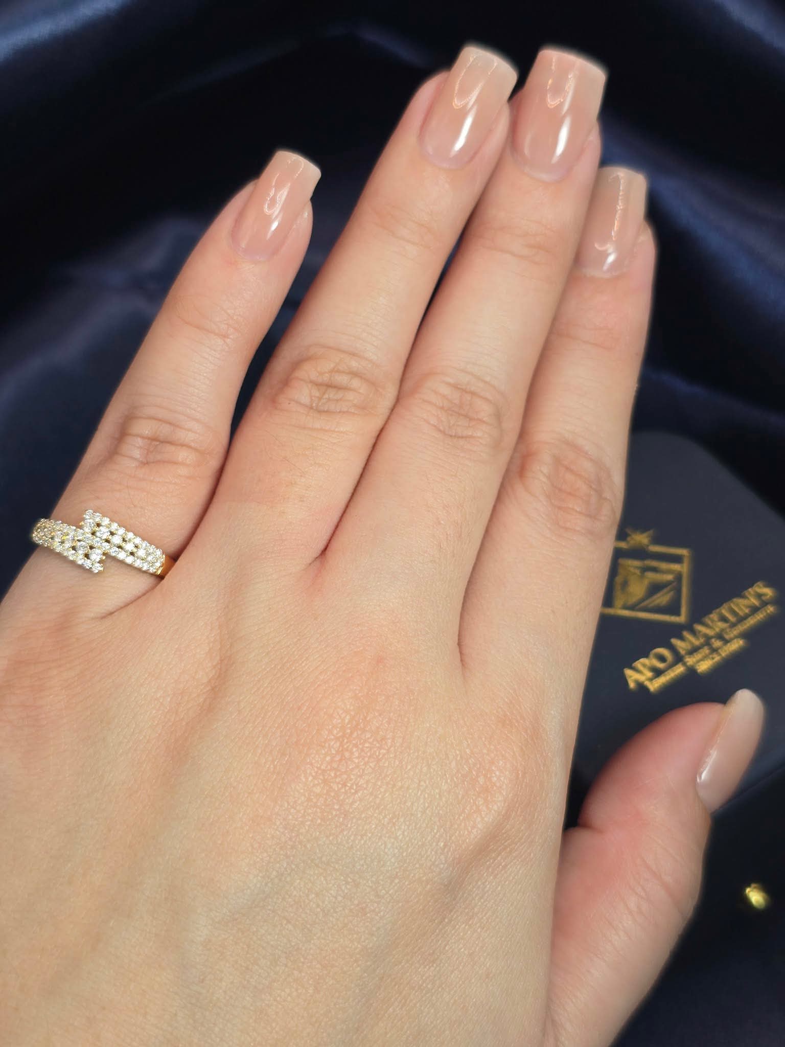 A close-up of a hand wearing a silver band ring with a geometric pattern of small clear stones, set against a dark navy blue satin background with a branded jewelry box.