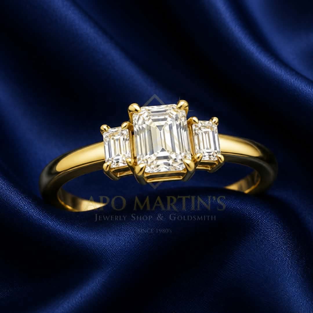 A yellow gold ring featuring a central emerald-cut diamond flanked by two baguette-cut diamonds, displayed on a dark blue satin fabric background. A watermark for 'APO MARTIN'S Jewelry Shop & Goldsmith' is visible in the lower center.