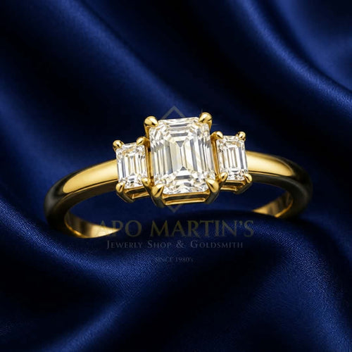 A yellow gold ring featuring a central emerald-cut diamond flanked by two baguette-cut diamonds, displayed on a dark blue satin fabric background. A watermark for 'APO MARTIN'S Jewelry Shop & Goldsmith' is visible in the lower center.