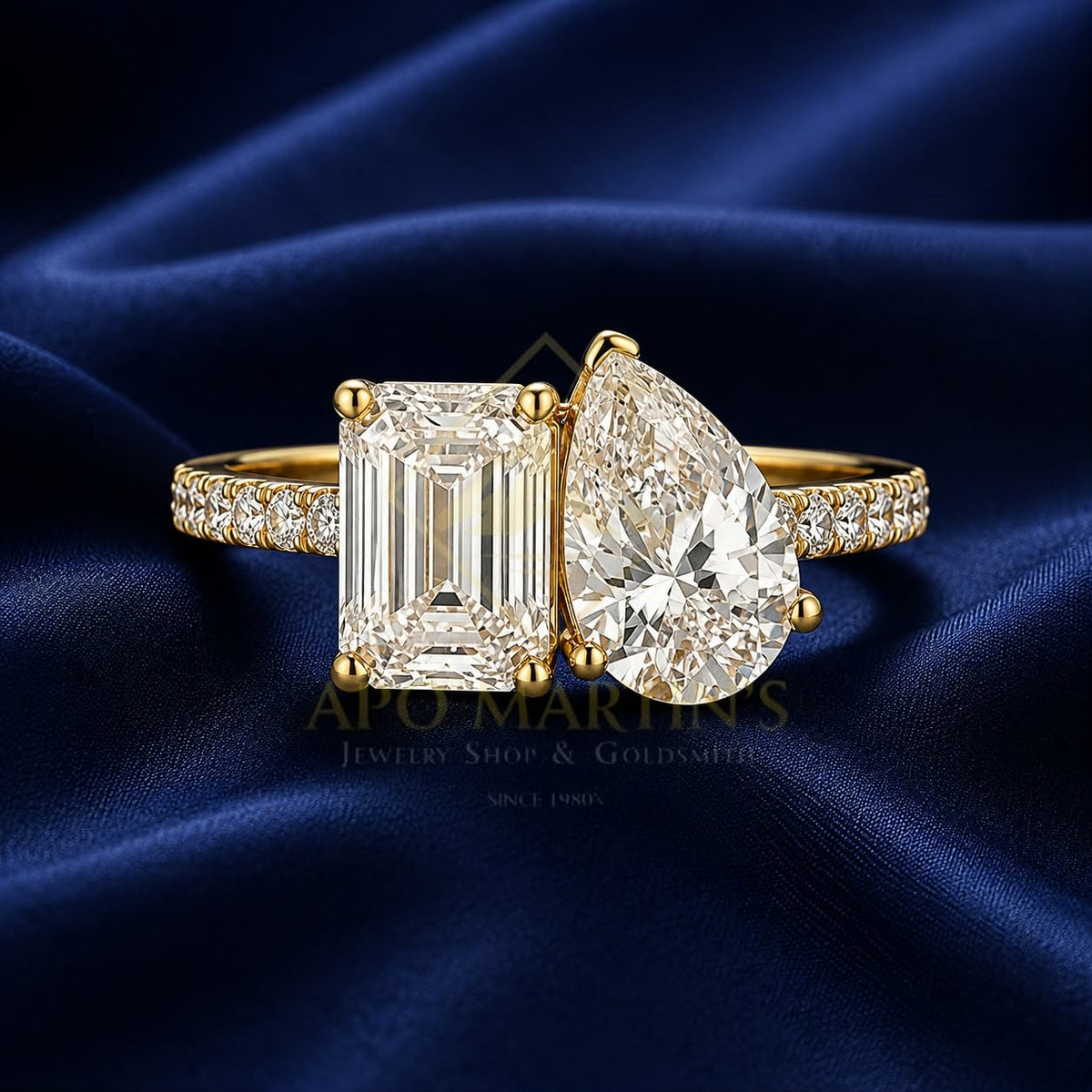 A yellow gold ring featuring a large emerald-cut diamond on one side and a pear-cut diamond on the other, with pavé-set diamonds along the band, displayed on a dark blue satin fabric background.