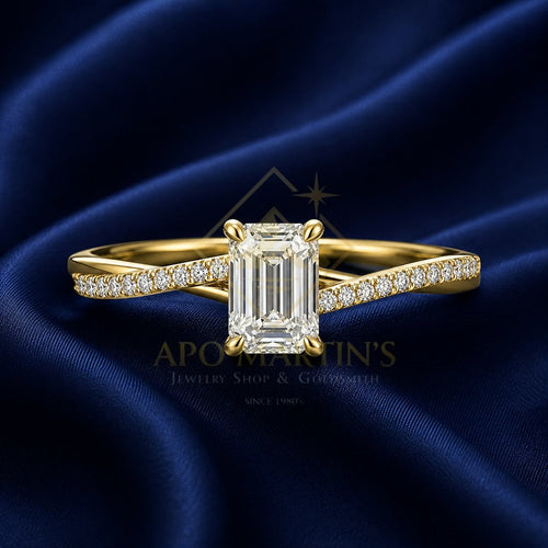 A yellow gold engagement ring with a large emerald-cut diamond center stone and pavé diamonds along the band, displayed on a draped dark blue satin fabric background. A watermark for 'APO MARTIN'S Jewelry Shop & Gemmology' is visible in the center.