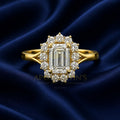 A yellow gold halo ring featuring a large emerald-cut center stone and surrounding round and baguette diamonds, displayed on a dark blue satin fabric background. A faint watermark for 'Apollo's Jewelry Shop & Goldsmith' is visible in the background.