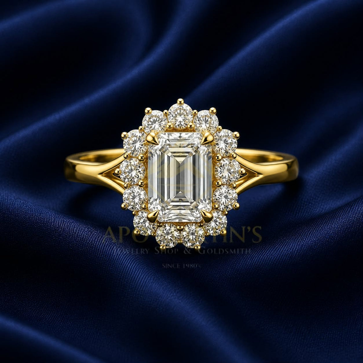 A yellow gold halo ring featuring a large emerald-cut center stone and surrounding round and baguette diamonds, displayed on a dark blue satin fabric background. A faint watermark for 'Apollo's Jewelry Shop & Goldsmith' is visible in the background.
