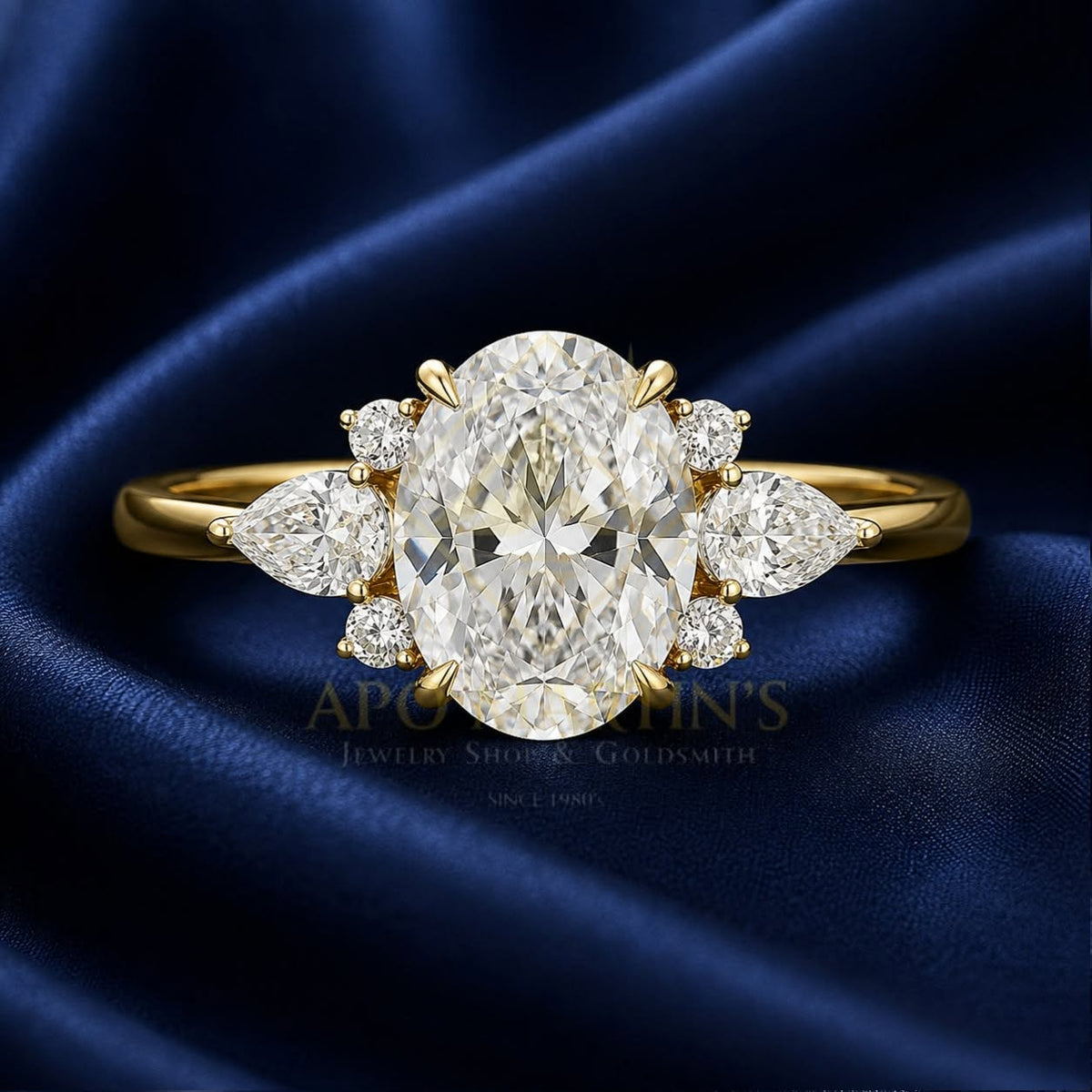 An oval-cut diamond engagement ring with marquise and round diamond accents on a yellow gold band, displayed on a dark blue satin fabric background. A watermark for 'Apollo Goldsmith's Jewelry Shop & Goldsmith' is visible across the center.