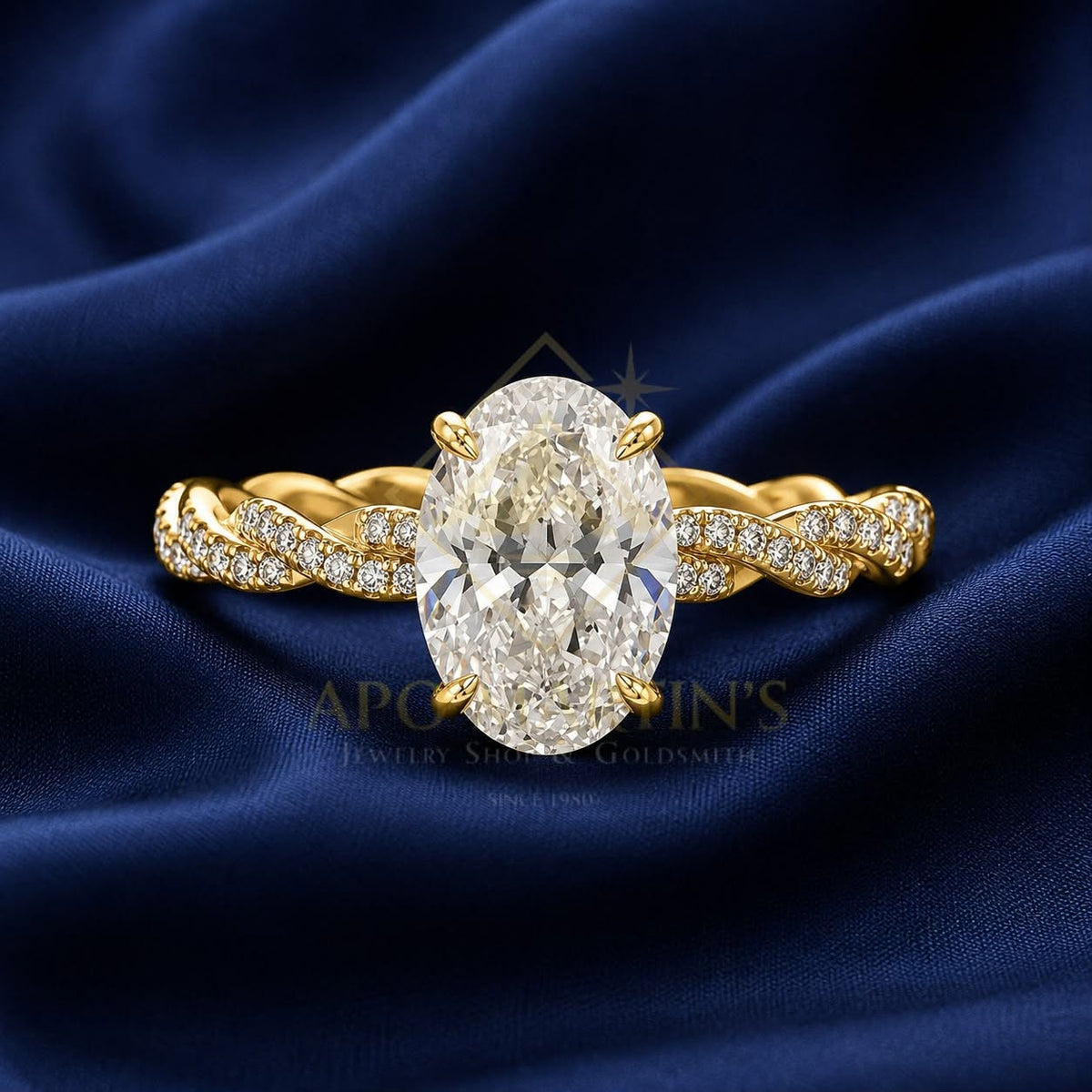 An oval diamond engagement ring with a yellow gold band featuring pavé-set diamonds, displayed on a draped navy blue satin fabric. A watermark for 'Apollo Goldsmith's' is visible in the center.