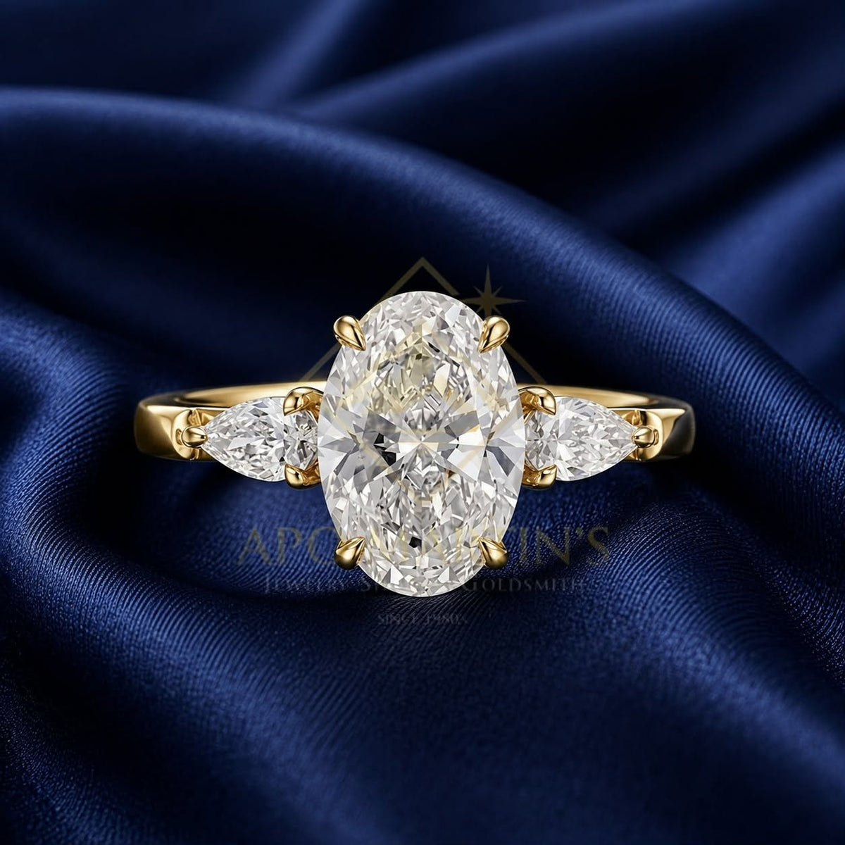 An oval diamond engagement ring with pear-cut side diamonds on a yellow gold band, displayed on a draped navy blue satin fabric background.